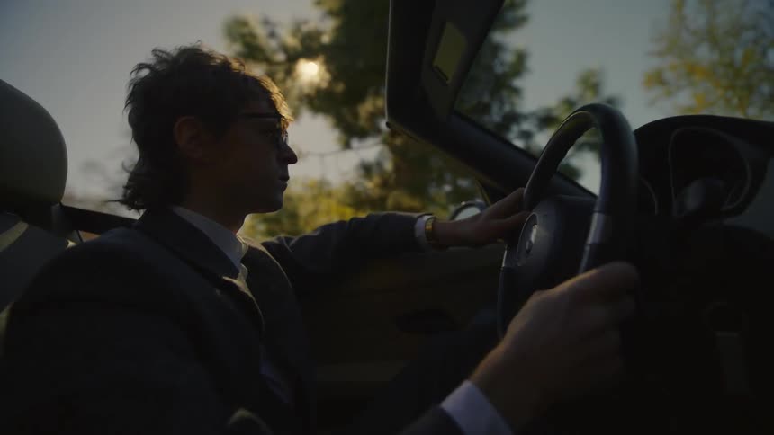 A man wearing sunglasses is driving a convertible car as the sun shines brightly in the distance