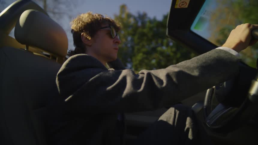 A man wearing sunglasses is driving a convertible car as rests his hand above the windshield