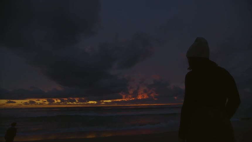 A young woman stands on a beach at sunset while another person stands nearby