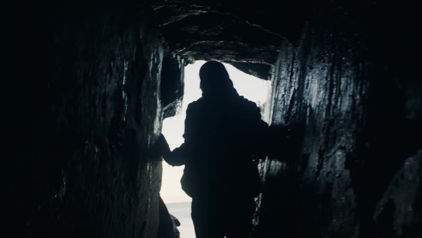 Person silhouetted against light from entrance walks down narrow tunnel with water dripping from its ceiling