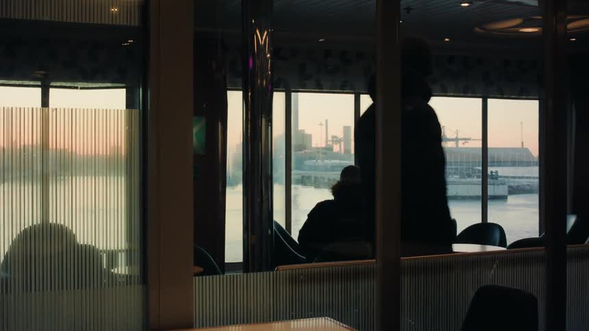 Man walks through restaurant as second person sits at table and looks out window at dock