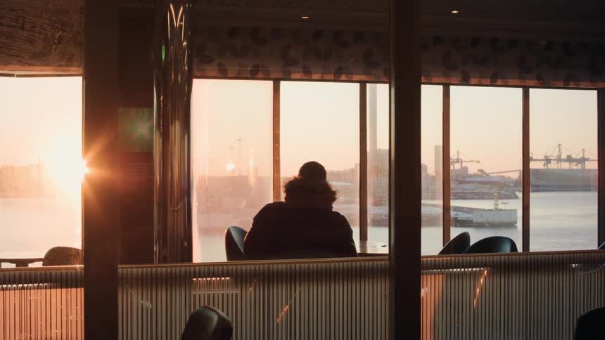 A person is sitting on a chair inside the ships lounge and watches the sunset as the ship passes a pier in the harbor