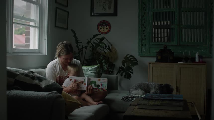 A mother and her infant son are sitting on a sofa in the livingroom reading a childrens book