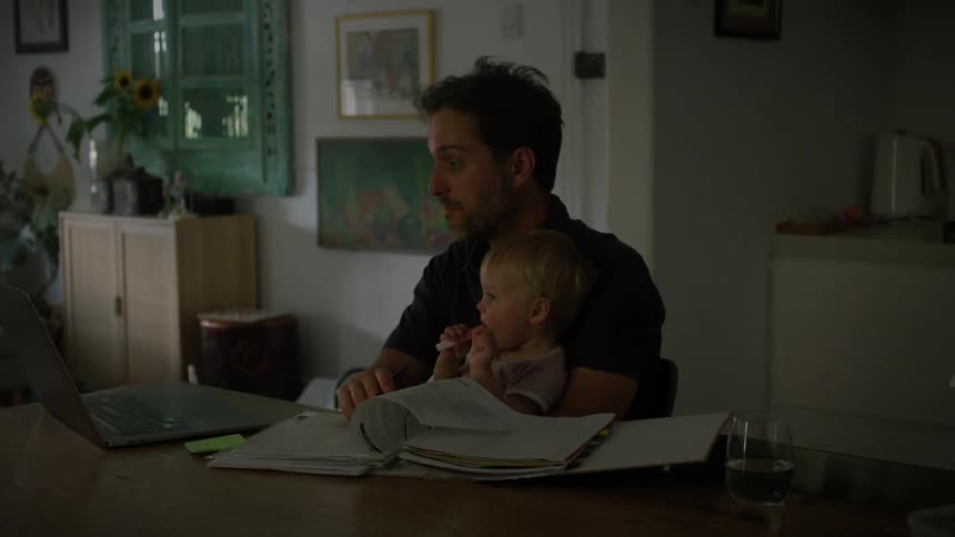 A father sits at the kitchen table while doing work and holding an infant