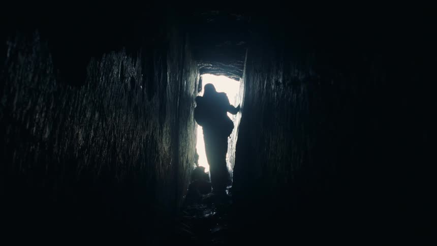 Backpacker is silhouetted against sunlight from entrance while walking into narrow tunnel