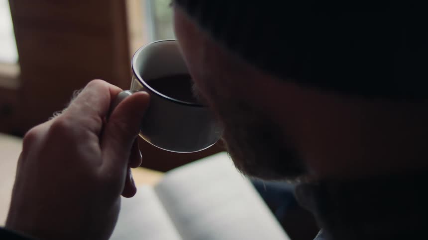 Older man sitting near window on a winter day sips a beverage while reading a book