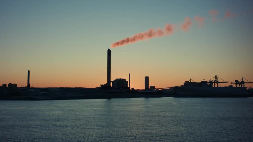 Waterfront manufacturing plant releases smoke while back lit by setting sun