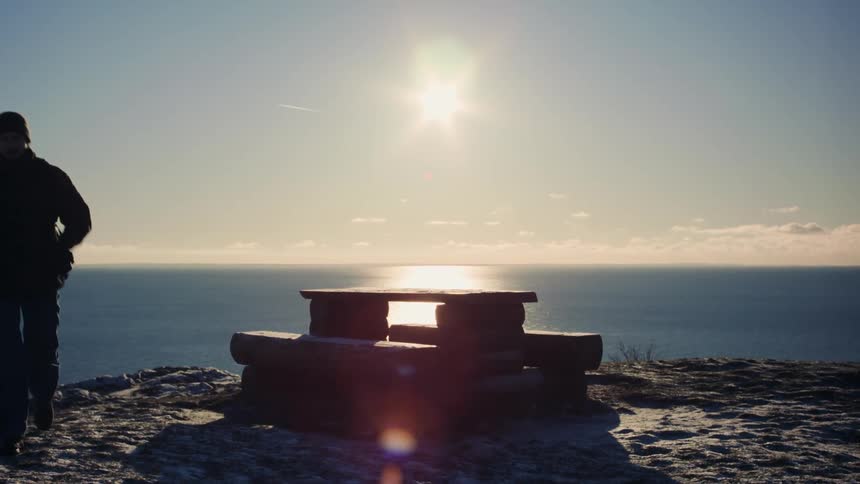 A male sitting on a table overlooking the ocean gets up and walks away