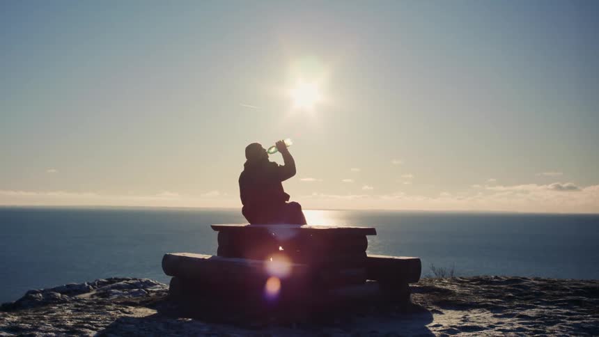 Man sitting on a structure at the peak of a seaside mountain drinking and enjoying the view with the sun on the horizon