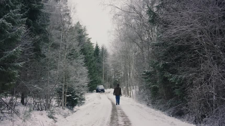 A person is walking on a snowy road towards a parked car surrounded by frosty trees