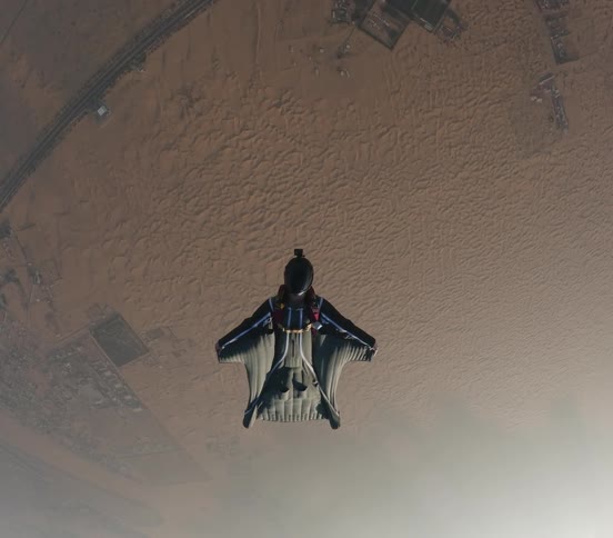 Person in wingsuit glides over buildings in arid terrain