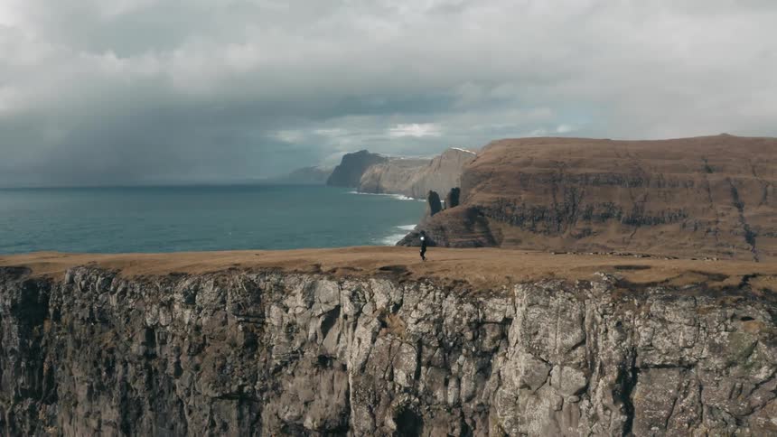 Person walks along the coastal cliff under cloudy sky near the sea