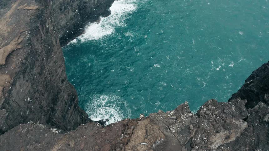 Snow flakes falling and starting to stick on oceanside cliffs as waves crash and foam below