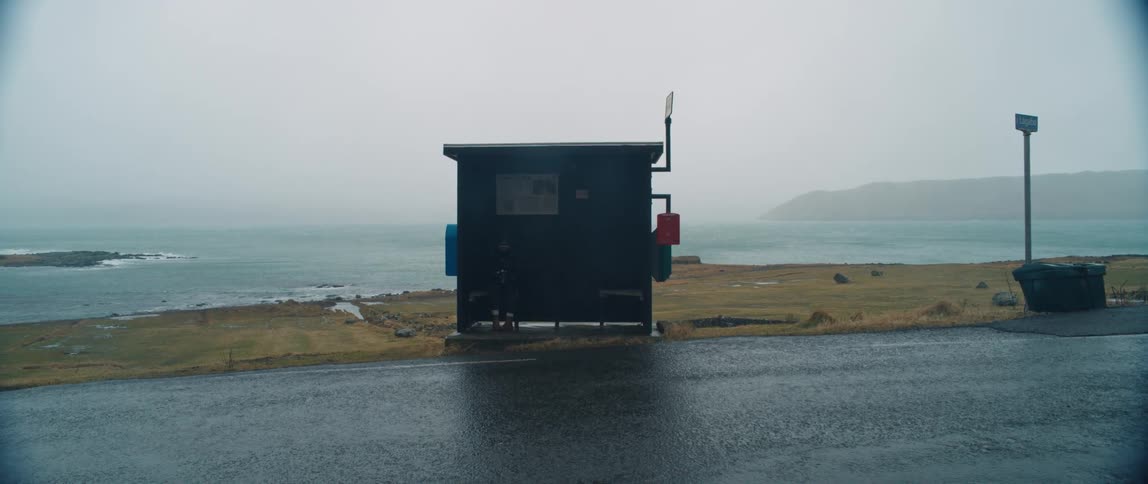 Person sits waiting at bus stop in field near coast on a foggy day