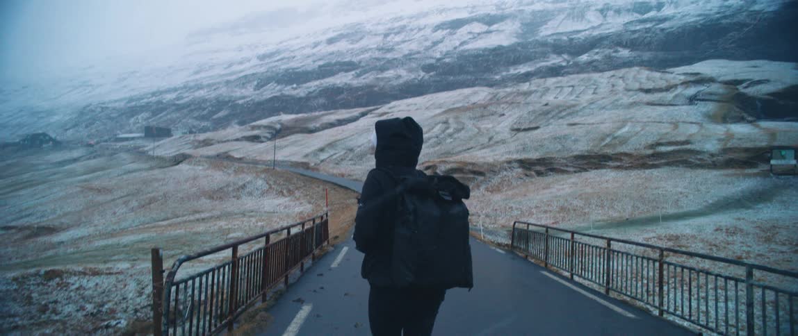 Woman with a backpack walking on a bridge amidst snowy mountains facing away