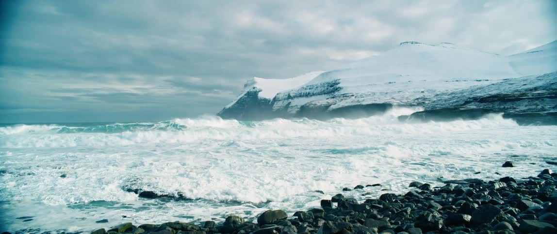 A rocky shoreline with turbulent waves and snowy mountains