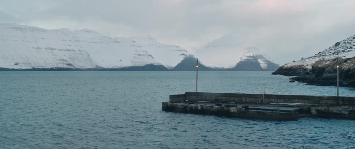 A pier juts into an ocean that has snow covered mountains behind it