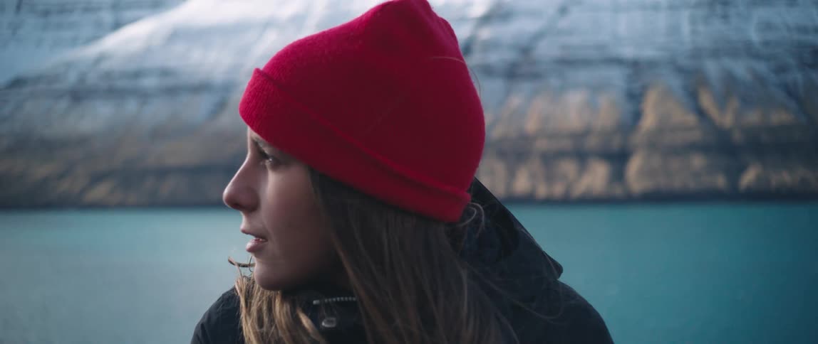 Woman with brown hair and a red wool hat stands by blue water near snowy mountain