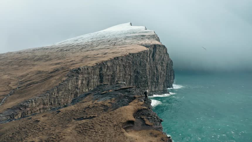 Aerial panorama of a person on the cliff of a promontory by the sea in winter on a cloudy day