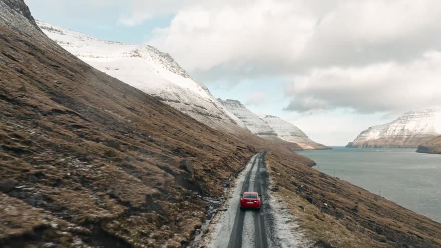 A car travels on a mountain road overlooking the coast with snow covered mountains in the distance