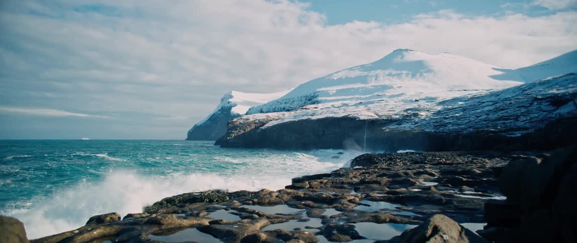 Sea waves crashing on a rocky shore and there is a huge mountain covered in snow