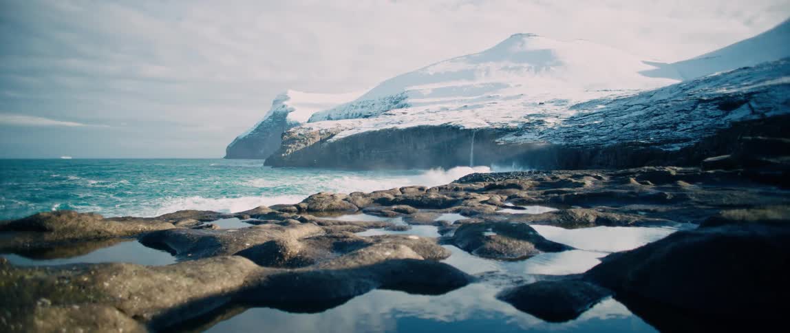 View of the sea with waves crashing against rocks in slow motion surrounded by snowcovered mountains on a cloudy day