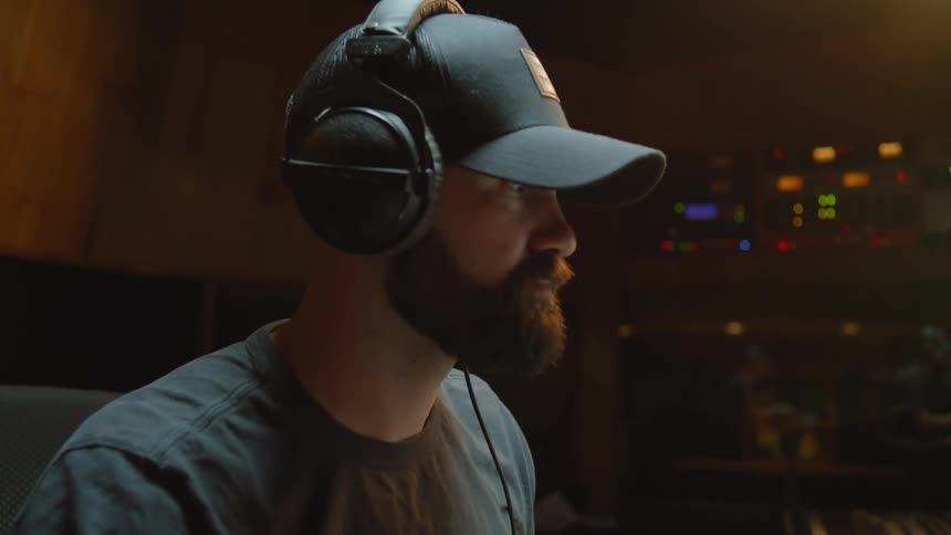 A man wearing a hat inside with headphones on bobbing his head to music while mixing it in a computer