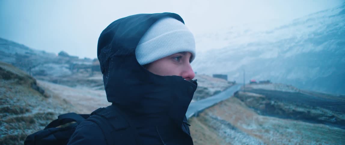 Person wearing beanie and jacket with hood stands outdoors near a mountain road in winter