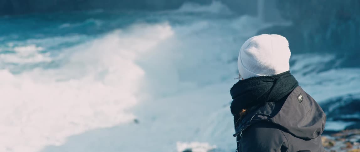 Woman with a white hat and black coat watches sea waves crashing against a cliff