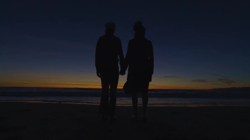 Silhouette of a couple one facing the sea and the other facing away with the sunrise on the horizon
