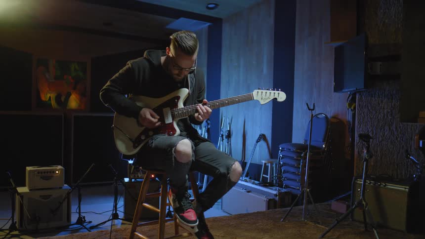 Man with glasses a black hoodie and ripped jeans plays a guitar sitting on a stool