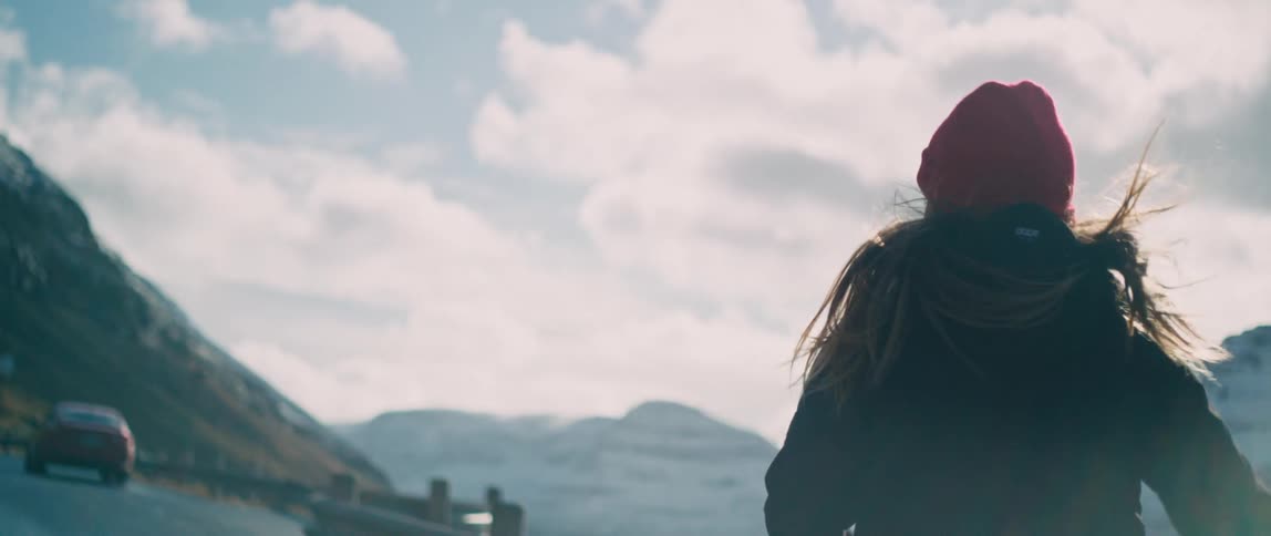 A woman looks back over her shoulder as she runs laughing toward a car on a mountain road overlook