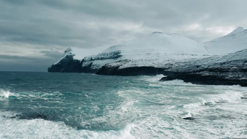 Aerial of ocean waves rolling into the shore with snow covered mountains in the background