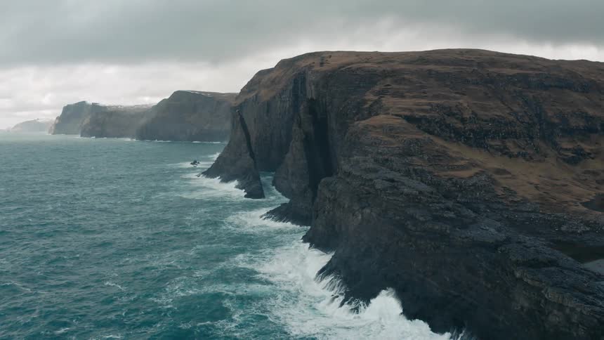 Waves splash against steep cliffs along the coast under a cloudy sky
