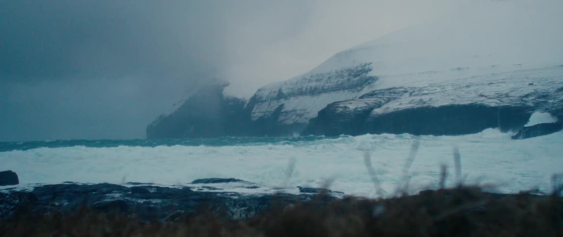 View of the sea with strong waves crashing against rocks surrounded by snowy mountains and promontory on a cloudy day