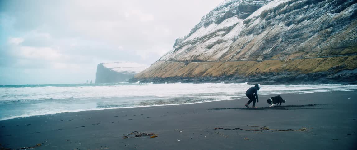 Man with a white hat black coat and pants plays with a dog on the black beach near the sea
