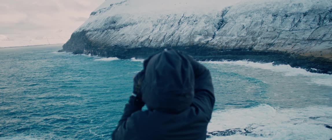 A person from behind is holding a camera and looking at the coast of a sea with mountains