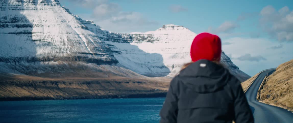Person wearing black jacket and red beanie walks along the road by the waterfront with snow covered mountains in the distance