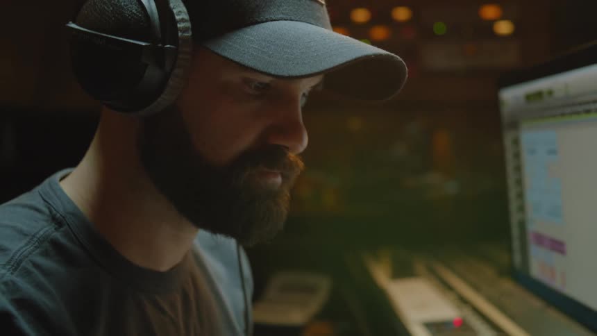A man wearing headphones and a cap is playing an electric piano and there is a screen in front of him