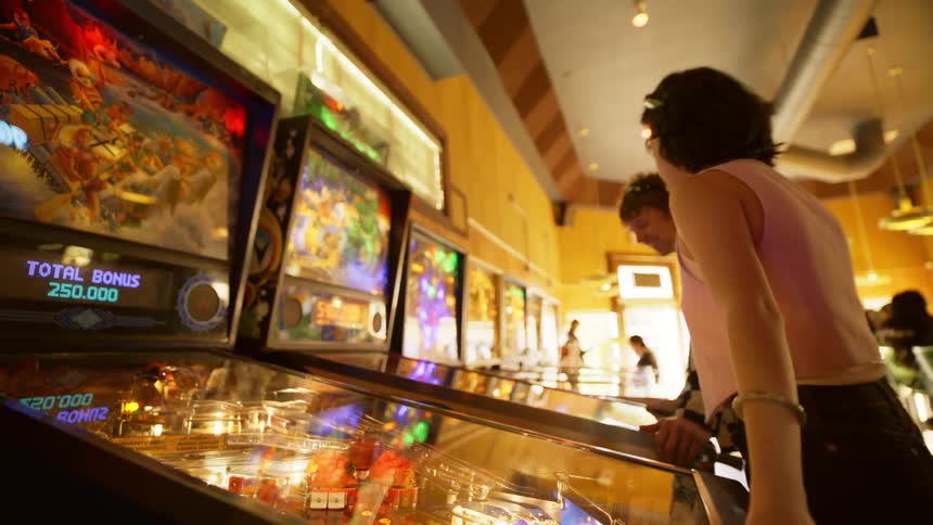 A teenage couple teases each other as they both play on separate pinball machines