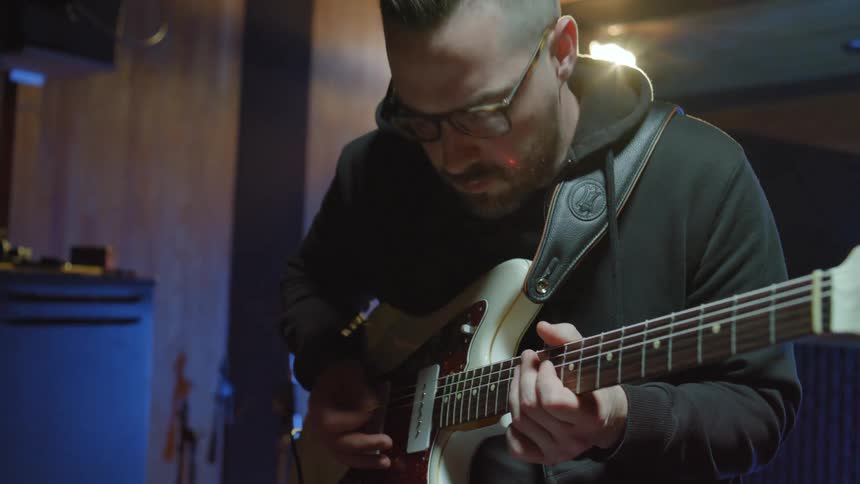 Man with glasses a beard and a black hoodie plays a guitar