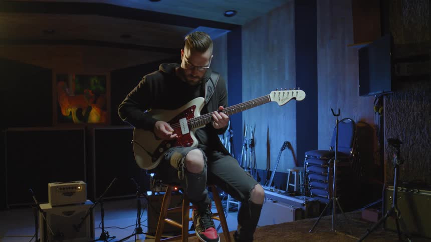 Man with glasses a beard a black hoodie and ripped jeans plays guitar sitting on a stool