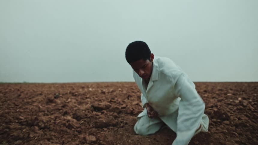 A man crouching digging a hole in the ground with his hands while holding a jewel with the other hand