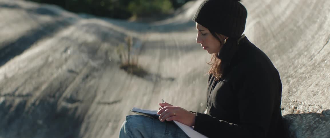 Woman wearing beanie sits outdoors sketching in notebook