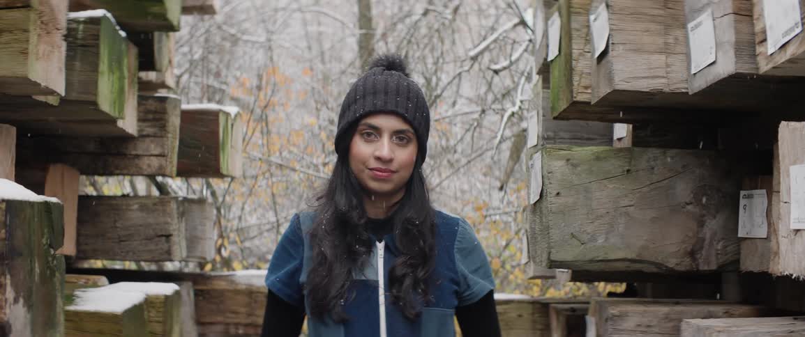Woman wearing a wooly hat and a fleece jacket stands among timber piles