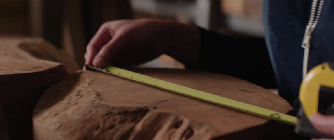 A person in a woodworking shop measures a wooden stump with measuring tape