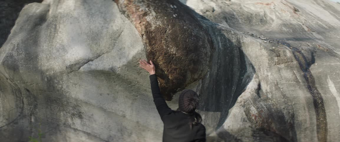 Person reaches left hand overhead and touches giant boulder outdoors