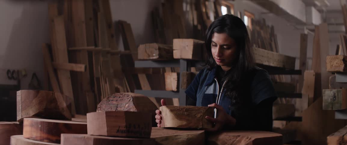 Female woodworker examines chunk of wood while standing among supplies in woodworking studio