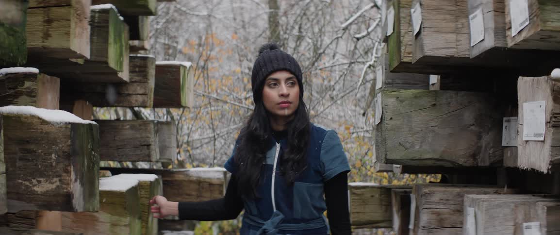 Woman wearing beanie walks outdoors between rows of stacked timber in the snow