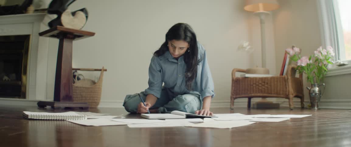 Female with dark hair sits on floor indoors drawing in sketchbook